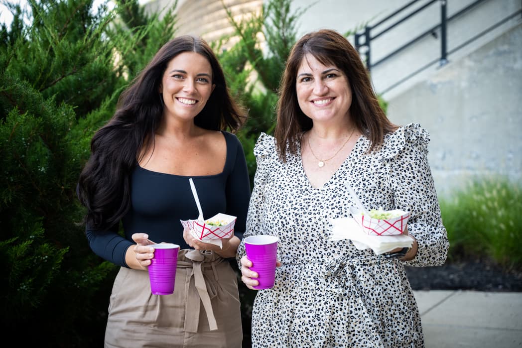 Two women holding food and drinks smiling at outdoor company cookout — Cincinnati event photographer K Dalton