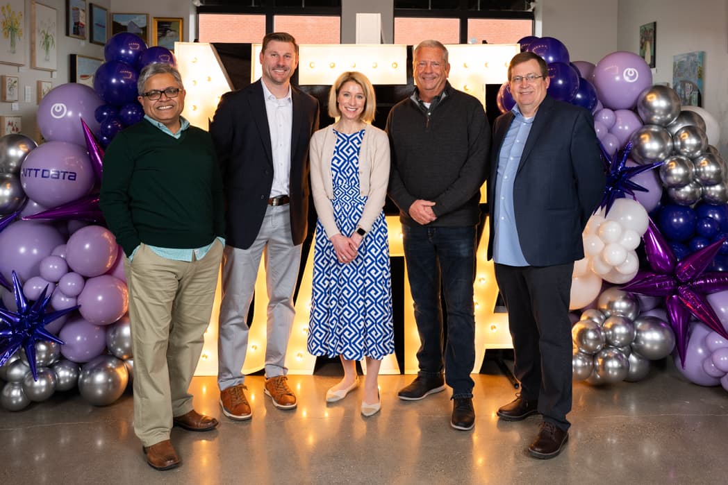 Five executives posing for group photo in front of illuminated NTT Data marquee letters with purple and silver balloon arrangements — Cincinnati event photographer K Dalton